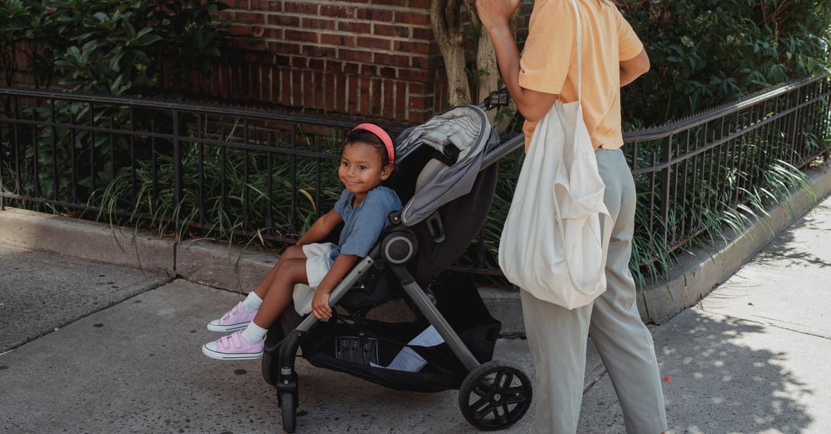 Crop Asian woman talking on smartphone while pushing baby stroller with little girl during walk on pavement in sunny city