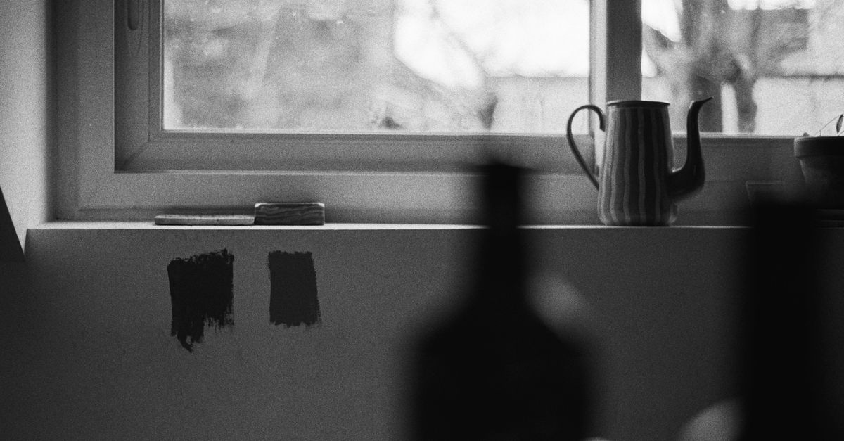 A black and white still life of a teapot on a windowsill with bottles and paint samples.