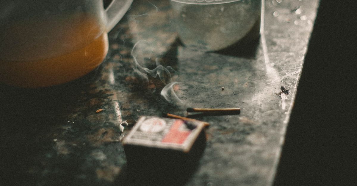 A moody still life depicting a burning matchstick next to orange juice and a plastic container on a rustic table.